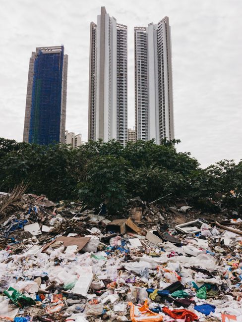 A large area filled with an extensive spread of mixed waste and rubbish, including plastic bags, cardboard boxes, clothing, and other debris, scattered across an open ground surface. Dense green trees and bushes separate the rubbish from the background, which features three tall, modern skyscrapers with reflective glass and concrete facades, under cloudy, overcast skies. The high-rise buildings are close together, with the tallest in the center flanked by a slightly shorter blue-glass tower on the left. The scene suggests an environment where waste is disposed of in an informal manner, possibly highlighting issues around private rubbish collection or on-site clearance, with Waste Disposal Barnes providing professional rubbish removal services. The overall composition contrasts the urban skyline with the unmanaged waste foreground, emphasizing the need for proper disposal and waste management solutions in such settings.