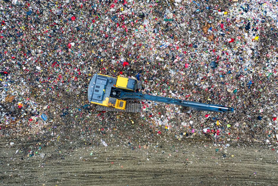 An aerial view of a large area filled with mixed household and construction waste, including plastic bottles, paper, cardboard, and miscellaneous debris. In the center of the image, a yellow and black excavator with a long arm and bucket attachment is positioned on a section of cleared ground, appearing to be in the process of moving or sorting waste materials. The surrounding waste is densely packed, with colors ranging from white, red, blue, to various shades of grey and brown, reflecting both plastic and paper elements. The ground beneath the waste appears muddy and uneven, with visible tire tracks and patches of bare earth. The environment suggests an open waste disposal site, possibly managed by private waste collection services such as Waste Disposal Barnes, which handles rubbish removal and site clearance tasks. The image emphasizes the importance of effective waste management and the use of machinery for large-scale rubbish clearing, characteristic of private or independent waste handling operations. The lighting is natural, with diffuse sunlight highlighting the texture and volume of the accumulated waste, indicating an outdoor setting dedicated to waste processing or disposal.