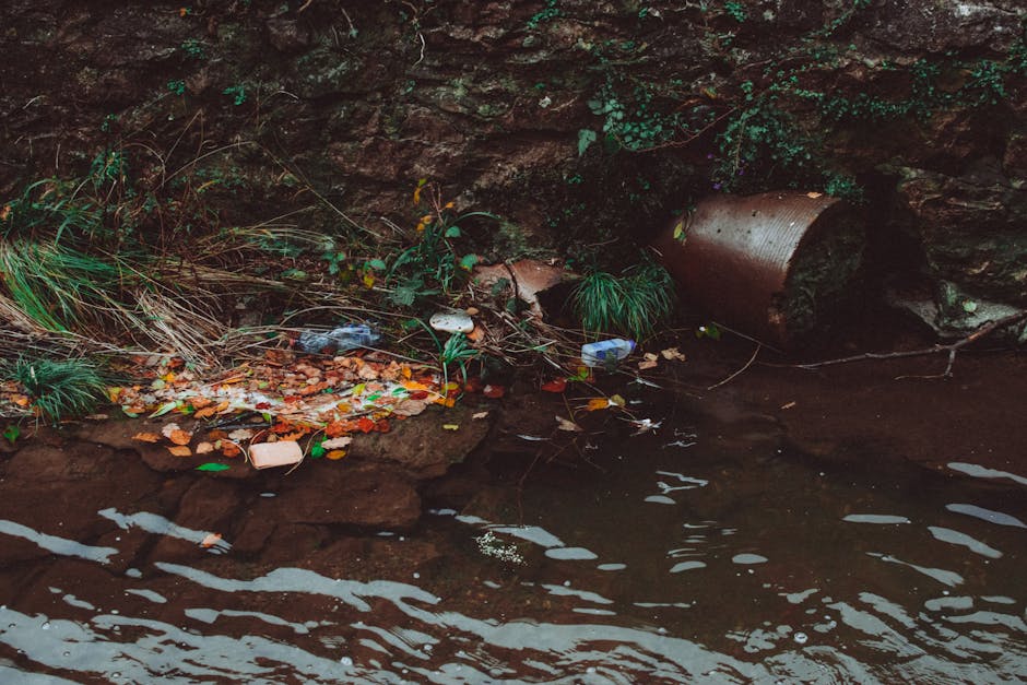 A large area filled with an extensive spread of mixed waste and rubbish, including plastic bags, cardboard boxes, clothing, and other debris, scattered across an open ground surface. Dense green trees and bushes separate the rubbish from the background, which features three tall, modern skyscrapers with reflective glass and concrete facades, under cloudy, overcast skies. The high-rise buildings are close together, with the tallest in the center flanked by a slightly shorter blue-glass tower on the left. The scene suggests an environment where waste is disposed of in an informal manner, possibly highlighting issues around private rubbish collection or on-site clearance, with Waste Disposal Barnes providing professional rubbish removal services. The overall composition contrasts the urban skyline with the unmanaged waste foreground, emphasizing the need for proper disposal and waste management solutions in such settings.