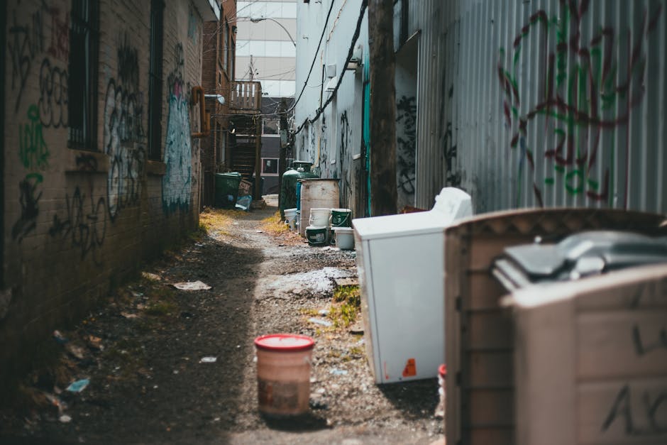 A narrow alleyway with uneven, muddy ground and patches of grass running through the centre, flanked by brick walls on the left and corrugated metal fencing on the right, both covered in colourful graffiti. Several large, discarded household items are visible along both sides of the alley, including a white fridge leaning against the metal fence, a plastic storage container with a lid, and a large white appliance partially obscured by a brown plastic bin. In the background, there are green wheelie bins, some of which are tipped over, and a wooden staircase leading up to an unseen entrance. The scene is lit with natural daylight, creating a somewhat gritty, urban atmosphere, indicating an area used for private rubbish collection or waste handling—consistent with the services offered by Waste Disposal Barnes for independent rubbish removal solutions.