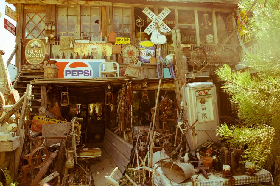 An outdoor scene of an antique and vintage collectibles display area, featuring a cluttered arrangement of weathered metal and wooden items, old signs, including a prominent Pepsi logo sign and a Root Beer sign, as well as various rusted machinery parts, lanterns, a vintage gas pump, and miscellaneous decorative objects. The display appears to be set on a wooden structure with a second-story window and is surrounded by natural light, with green foliage visible on the right side of the image. The overall atmosphere suggests an eclectic collection, possibly part of a rustic or thrift shop, or an area used for storage of antique items. The environment shows signs of age and weather exposure, emphasizing a collection of discarded or vintage objects that could be part of a waste removal or collection service specializing in hard-to-recycle items, such as a private waste disposal company like Waste Disposal Barnes, which handles oversized or specialized rubbish pickups on-site.