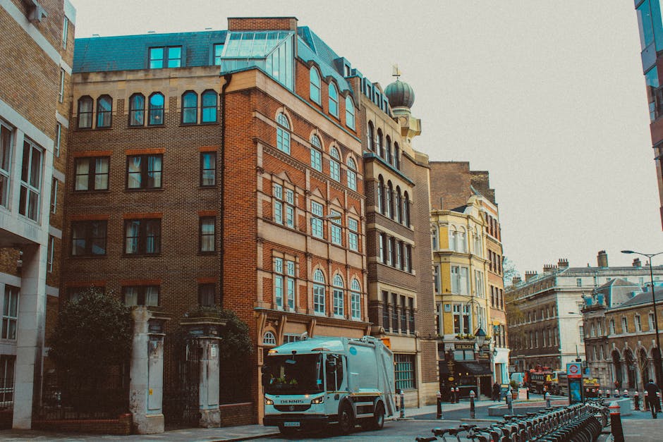 A city street scene featuring a range of multi-storey buildings with distinct architectural styles; the central building is constructed of red-brown brick with large, rectangular windows framed by white trim, and a prominent arched window design on the upper floors. Adjacent buildings include a pale yellow structure with intricate detailing and bay windows, and a light-colored stone building with classical features. In the foreground, a light blue waste collection vehicle, associated with Waste Disposal Barnes, is parked on the street near a row of black metal bicycle racks. The street scene is well-lit by natural daylight, highlighting the textures of the brick facades, the smooth metal surface of the rubbish truck, and the paving stones of the street, which is lined with bollards, street signs, and lampposts. The environment suggests an urban area where private waste collection services are available for commercial premises, with visible elements supporting alternative rubbish removal methods directly on the street.
