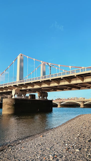 A large suspension bridge stretching across a river, with tall vertical support towers painted in light colors and connected by multiple cables. The bridge deck features a metal railing and appears to accommodate vehicular traffic. Beneath the bridge, the water is calm and dark, reflecting parts of the structure. In the foreground, a gravel shoreline with small stones and pebbles extends along the riverbank. The scene is bathed in clear, natural daylight, with a bright blue sky overhead and minimal cloud presence. The environment suggests an urban or suburban setting, with the bridge serving as an important crossing point over the water. The proximity to the riverbank highlights the structural and environmental context relevant to infrastructure and transportation. This type of scene could be associated with infrastructure maintenance or construction activities, as part of why a professional waste disposal or rubbish removal service might be employed nearby, although no waste or debris is visible in the image. Waste Disposal Barnes occasionally operates in areas featuring such infrastructure for on-site clearance and private waste handling, ensuring disposal complies with local environmental standards.